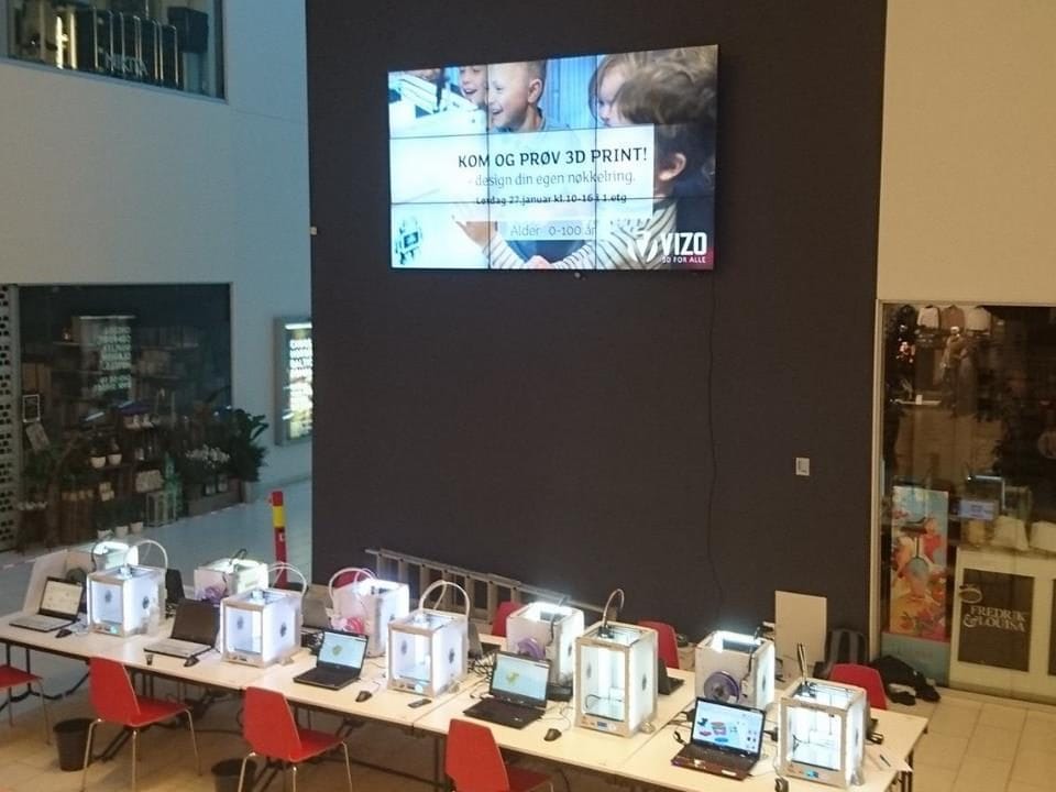 Stor DigiNoti-skjerm på en mørk vegg over en utstilling av 3D-printere
