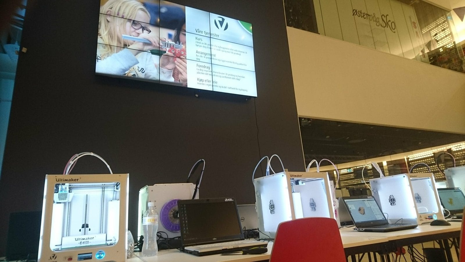 Veggmontert infoskjerm over et bord med 3D-printere og bærbare PC-er